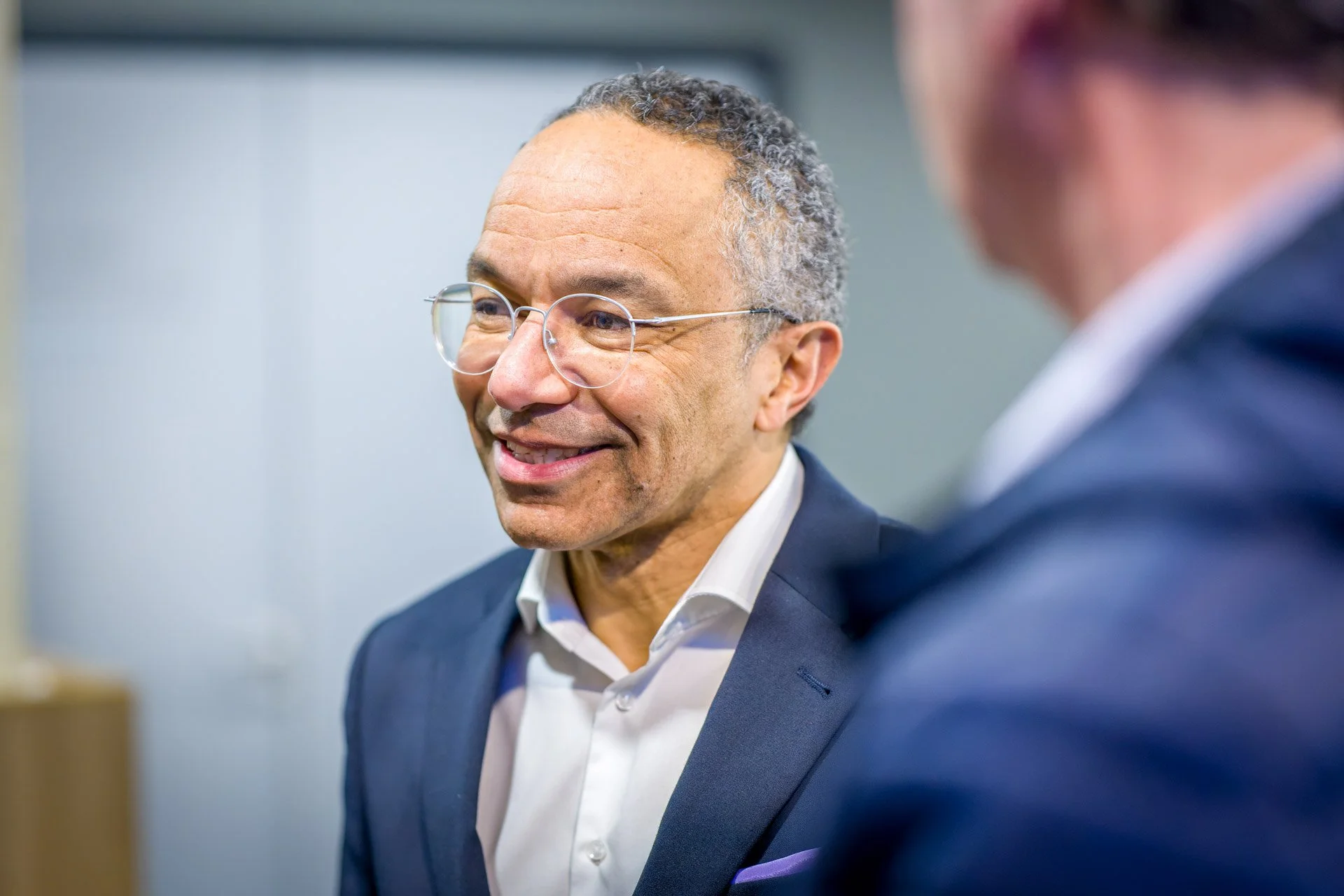 Un homme souriant avec des lunettes et une chemise blanche, discutant avec une autre personne floue au premier plan.