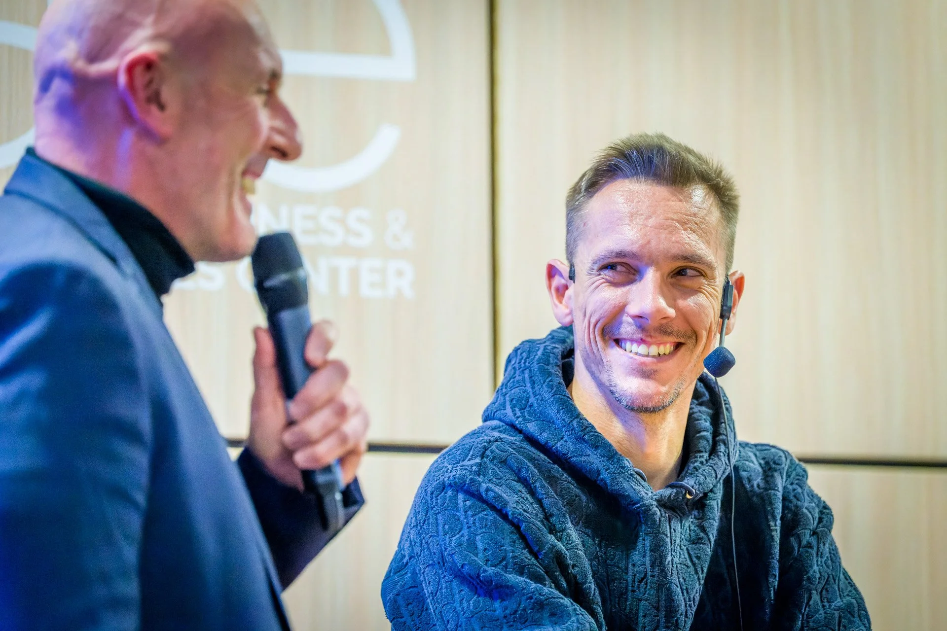 Deux hommes souriants lors d'un événement en intérieur, l'un tenant un microphone, l'autre portant un micro-casque.
