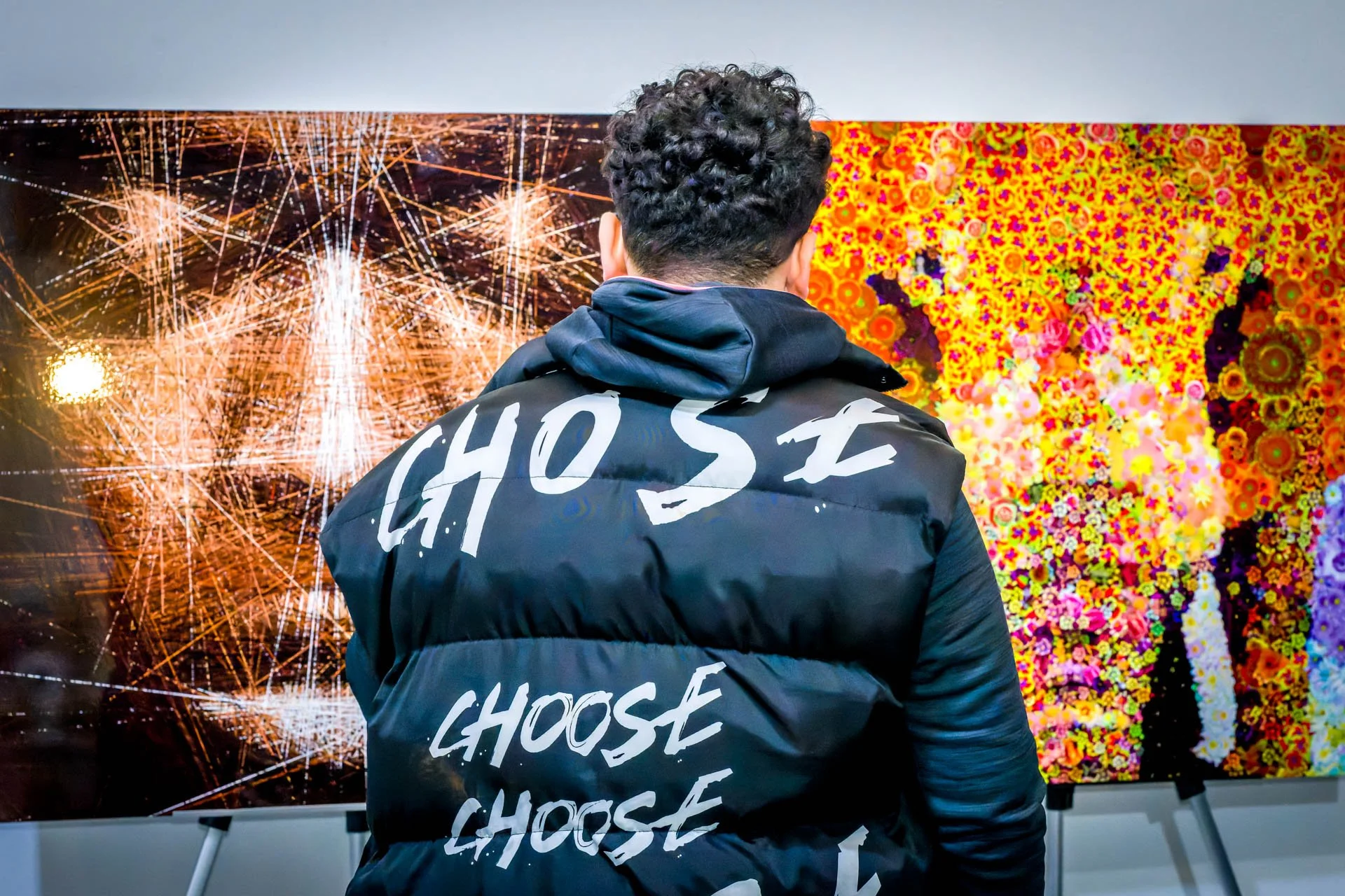 un événement organisé dans une galerie d'art à Bruxelles. Un homme de dos portant une veste avec l'inscription "CHOOSE" devant une œuvre d'art abstraite colorée sur un mur.